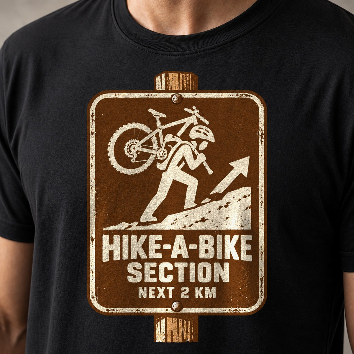 Hike-A-Bike Section Next 2 KM mountain bike t-shirt in Black — brown park-style trail sign of a rider carrying a bike uphill — close-up of the print